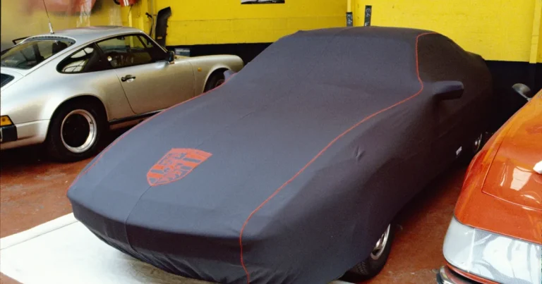 Porsche remisée sous une housse grise à passepoil orange et écusson brodé, dans un garage à côté d'une 911